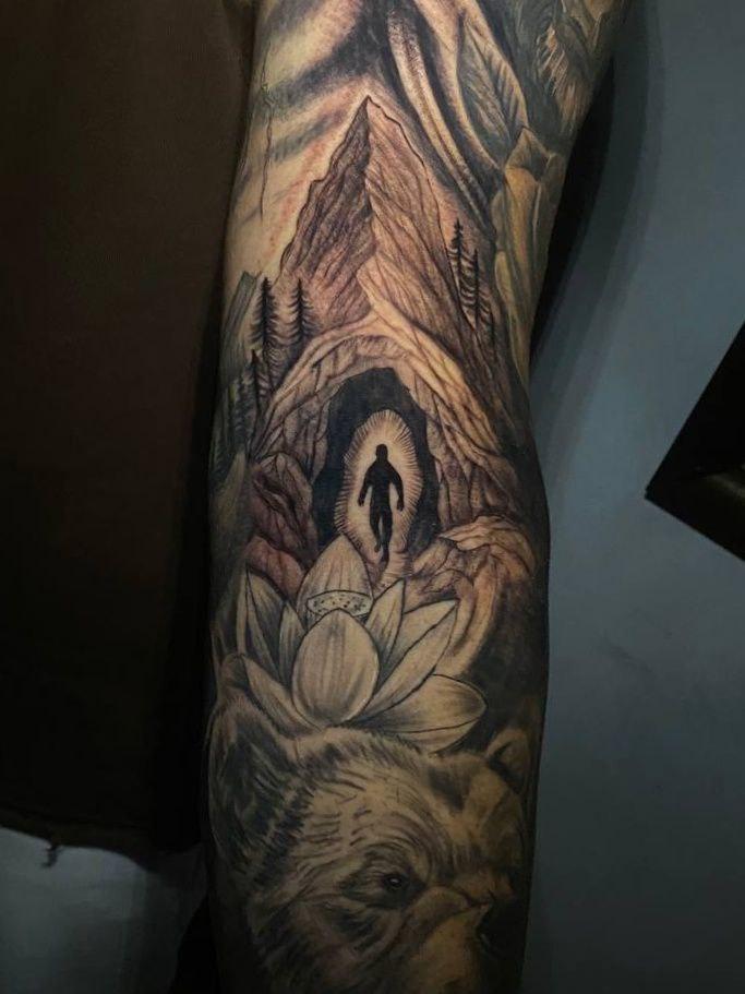Tatuaje realista con montaña, cueva luminosa y flor de loto en el brazo